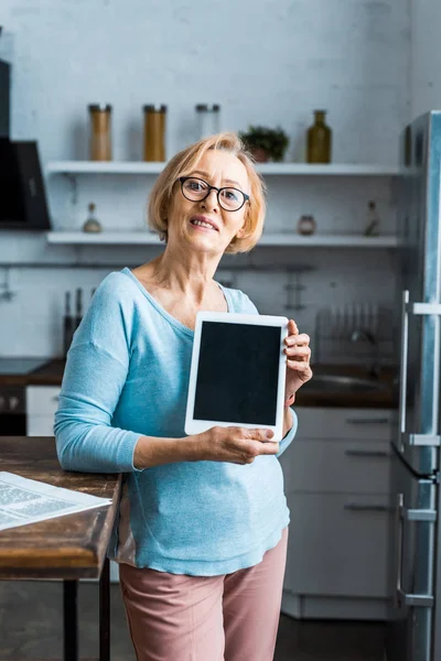 Seniorin mit Brille blickt in Kamera und präsentiert digitales Tablet mit leerem Bildschirm in Küche — Stockfoto