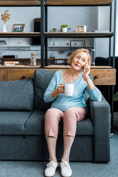 Пожилая женщина с чашкой кофе сидит на диване и разговаривает на смартфоне в гостиной — стоковое фото