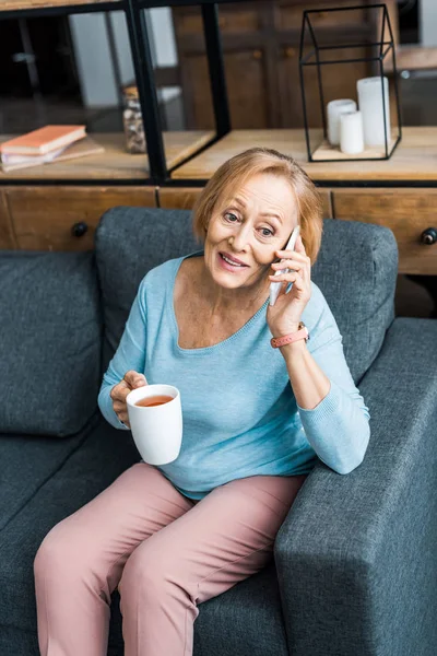 Felice donna anziana con una tazza di caffè seduta sul divano e parlare su smartphone in soggiorno — Foto stock