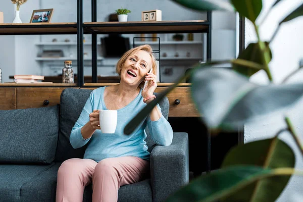 Rire femme âgée avec tasse de café assis sur le canapé et parler sur smartphone dans le salon — Photo de stock
