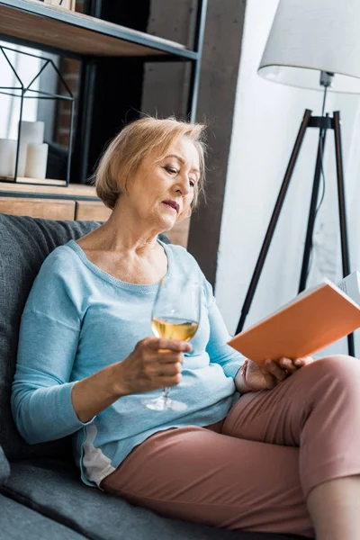 Senior woman sitting on couch, holding wine glass and reading book at home — Stock Photo