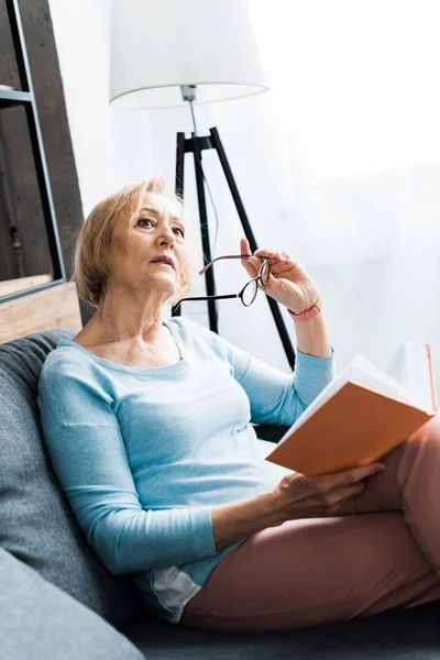 Nachdenkliche Seniorin sitzt auf Couch, hält Brille und schaut weg, während sie zu Hause Buch liest — Stockfoto