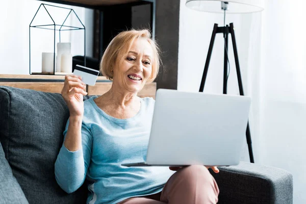 Счастливая пожилая женщина сидит, держа кредитную карту и используя ноутбук, делая покупки в Интернете дома — стоковое фото