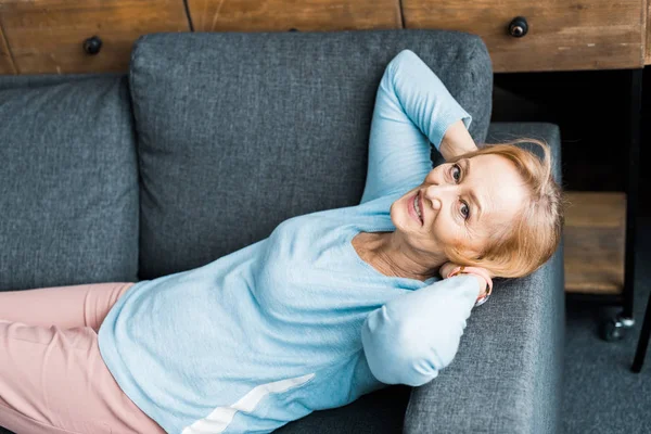 Femme âgée souriante avec les mains sur la tête regardant la caméra, couchée et reposant sur le canapé — Photo de stock