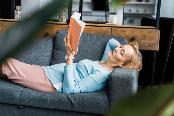 Селективное внимание пожилой женщины, лежащей на диване и читающей книгу дома — стоковое фото