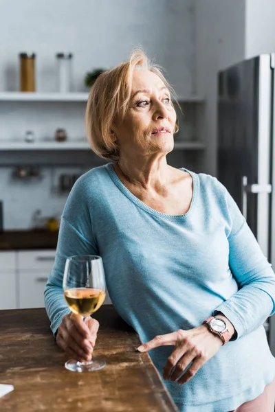 Solitaire femme âgée tenant verre de vin dans la cuisine et détournant les yeux — Photo de stock