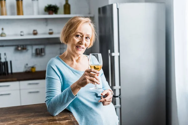 Donna anziana sorridente guardando la fotocamera e tenendo il bicchiere di vino in cucina con spazio copia — Foto stock