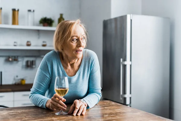 Одинокая пожилая женщина держит бокал вина на кухне и смотрит в сторону — стоковое фото