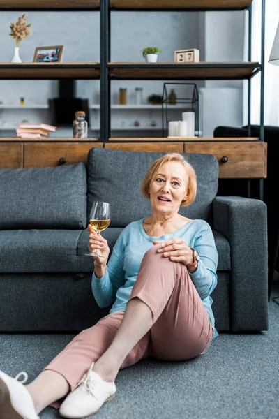 Donna anziana seduta, guardando la fotocamera e tenendo il bicchiere di vino in soggiorno — Foto stock