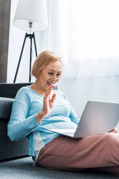 Lächelnde Seniorin winkt mit der Hand, während sie zu Hause Videotelefonie auf dem Laptop hat — Stockfoto