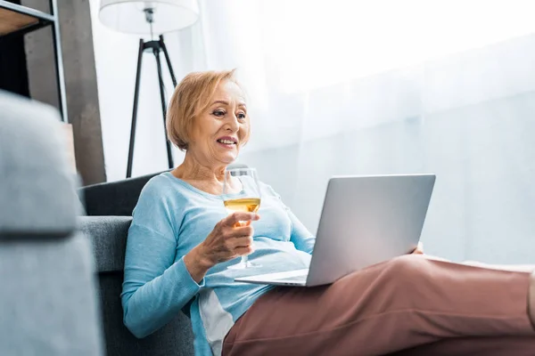 Heureux senior femme tenant verre de vin tout en ayant appel vidéo sur ordinateur portable à la maison — Photo de stock