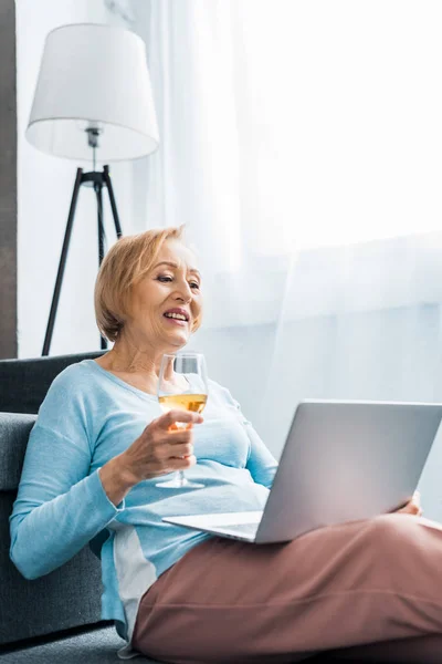 Усміхнена старша жінка тримає келих вина під час відеодзвінка на ноутбук вдома — стокове фото