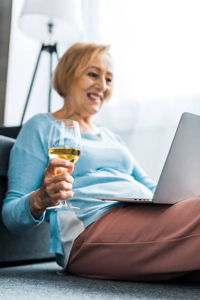 Souriant femme âgée tenant verre de vin tout en ayant appel vidéo sur ordinateur portable à la maison — Photo de stock