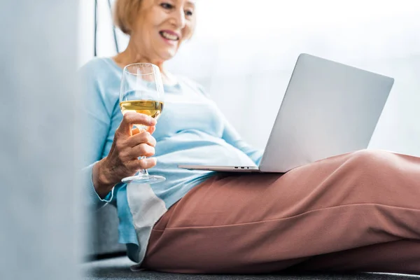 Senior femme tenant verre de vin tout en ayant appel vidéo sur ordinateur portable à la maison — Photo de stock