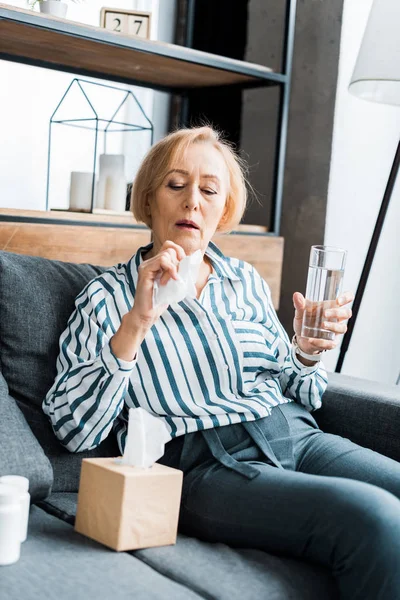 Sick senior woman suffering from cold and holding glass of water with tissue while sitting at home — Stock Photo