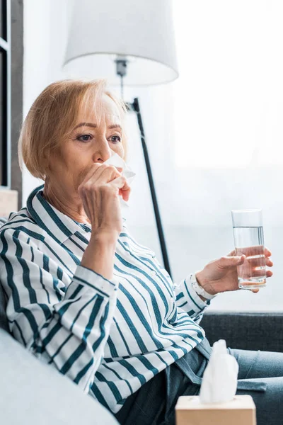 Sick senior woman with runny nose suffering from cold and holding glass of water at home — Stock Photo