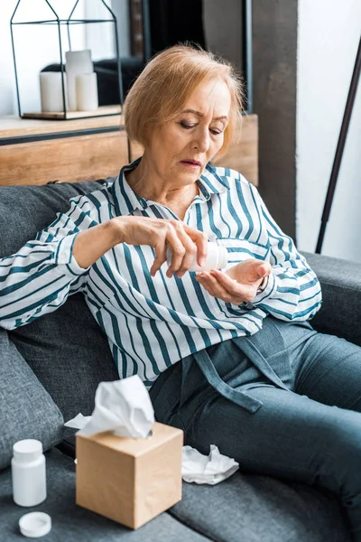 Sick senior woman sitting on couch with tissue box and taking pills at home — Stock Photo