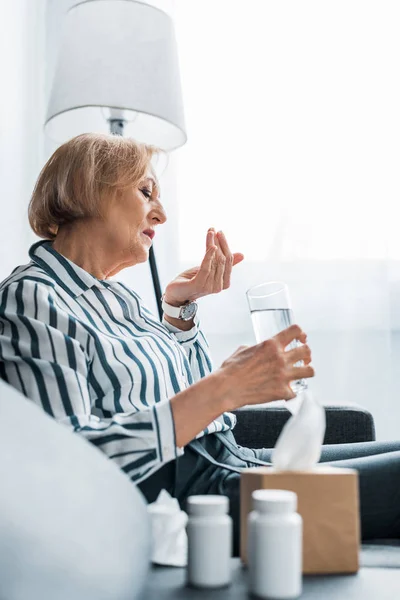 Enfoque selectivo de la mujer mayor enferma tomando pastillas y sosteniendo un vaso de agua en casa - foto de stock