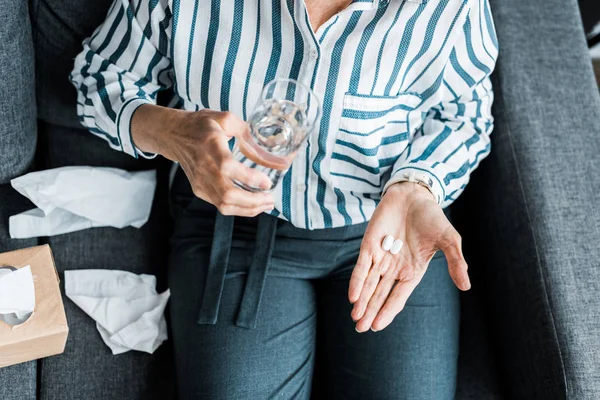 Cropped view of sick senior woman sitting on couch with pills and glass of water at home — Stock Photo