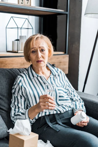 Sick senior woman looking at camera and holding bottle of medication with glass of water — Stock Photo