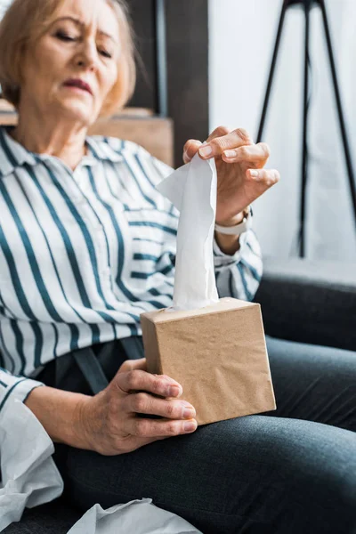 Sick senior woman suffering from cold and reaching for tissue while sitting at home — Stock Photo