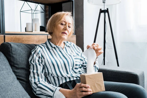 Sick senior woman sitting on couch, suffering from cold and reaching for tissue — Stock Photo