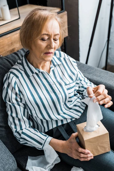 Donna anziana malata che soffre di raffreddore e cerca tessuto mentre è seduta a casa — Foto stock