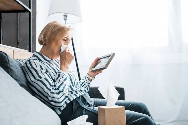 Mujer mayor deprimida llorando y limpiando la cara de las lágrimas con tejido mientras mira el marco de la imagen - foto de stock