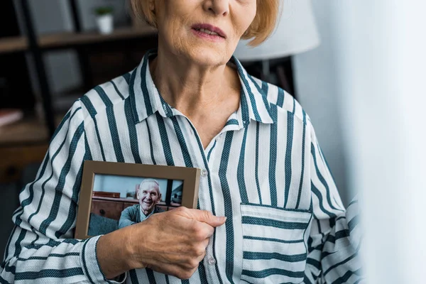 Частичный вид пожилой женщины, держащей рамку с мужчиной на фотографии дома — стоковое фото