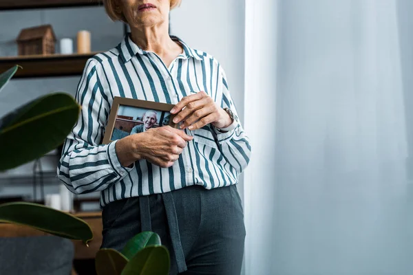 Частичный вид пожилой женщины, держащей рамку с мужчиной на фотографии дома с копировальным пространством — стоковое фото