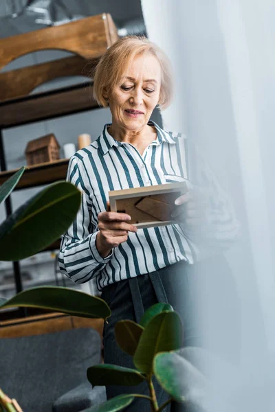 Foco seletivo da mulher idosa olhando para moldura de imagem em casa — Fotografia de Stock