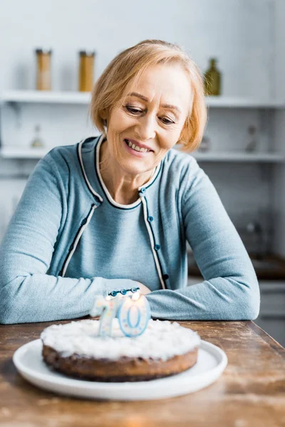 Пожилая женщина сидит за столом и смотрит на торт с табличкой 