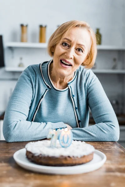 Donna anziana seduta a tavola e guardando la torta della macchina fotografica con il segno degli anni '70 durante la festa di compleanno a casa — Foto stock