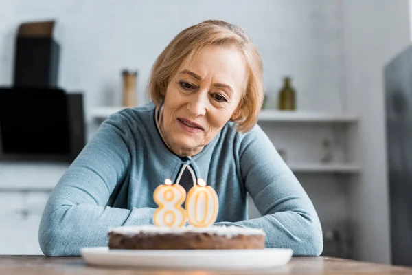 Donna anziana sola seduta a tavola e che guarda la torta con il cartello degli anni '80 durante la festa di compleanno a casa — Foto stock
