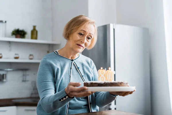 Femme âgée solitaire tenant gâteau avec '80' signe sur le dessus pendant la célébration d'anniversaire — Photo de stock