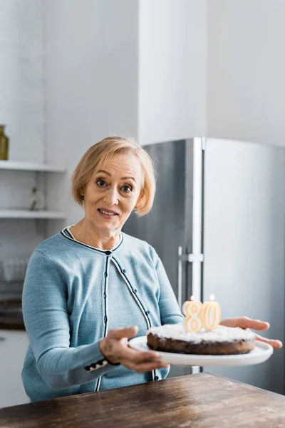 Пожилая женщина смотрит в камеру и держит торт с надписью 