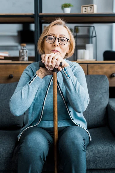 Turbato donna anziana in occhiali seduto sul divano, guardando la fotocamera e appoggiandosi sul bastone da passeggio in soggiorno — Foto stock