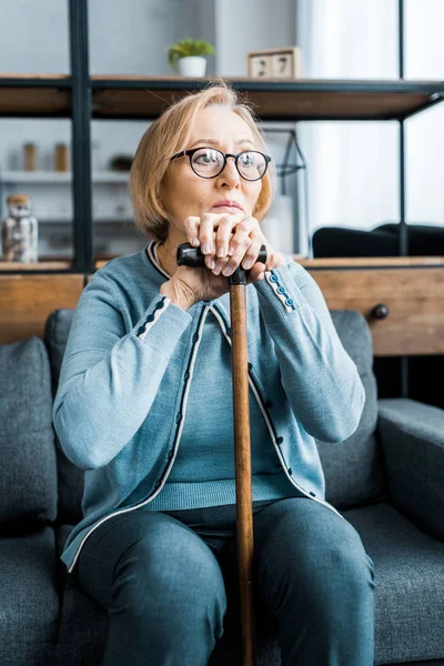 Turbato donna anziana in occhiali seduto sul divano e appoggiato sul bastone da passeggio in soggiorno — Foto stock