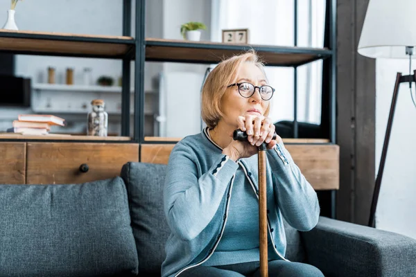 Verärgerte Seniorin mit Brille sitzt auf Couch und stützt sich im Wohnzimmer auf Gehstock — Stockfoto