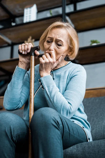 Depressed senior woman holding walking stick, covering mouth with hand and crying at home — Stock Photo