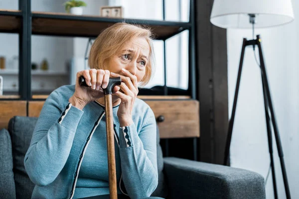 Upset senior woman holding walking stick, covering mouth with hand and crying at home with copy space — Stock Photo