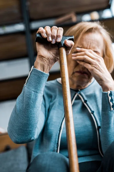 Mujer mayor molesta sosteniendo bastón, cubriendo la cara con la mano y llorando en casa - foto de stock