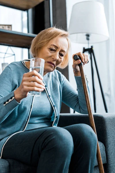 Sad senior woman sitting with walking stick and glass of water at home — Stock Photo