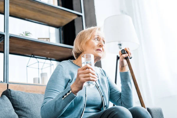 Sad senior woman sitting with walking stick and glass of water at home — Stock Photo