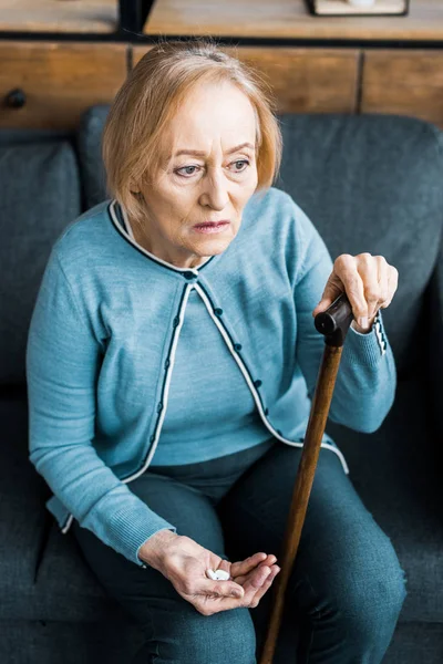 Femme âgée malade avec bâton de marche assis sur le canapé et tenant la médecine dans la main — Photo de stock