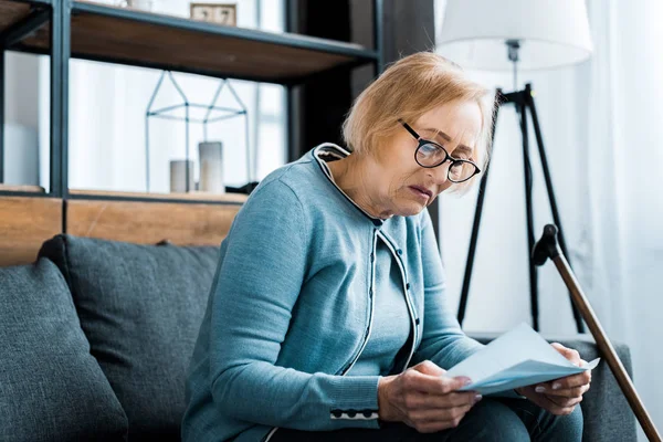 Senior woman in glasses sitting on couch and holding tax form at home — Stock Photo