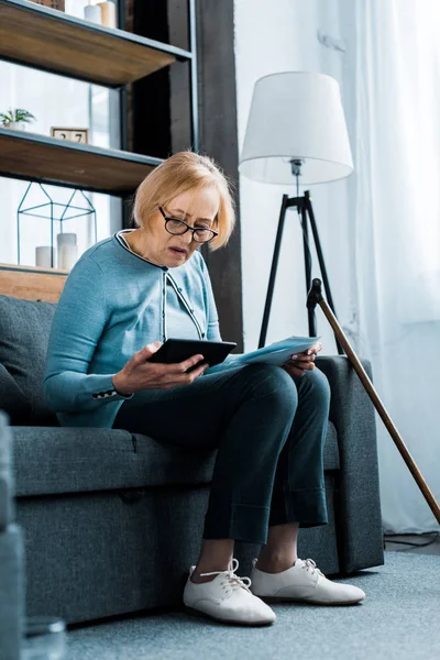 Seniorin sitzt mit Taschenrechner auf Couch und zählt Rechnungen im Wohnzimmer — Stockfoto