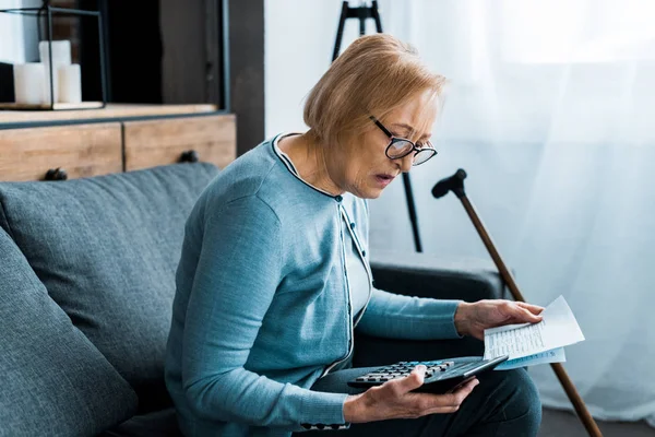 Mulher idosa sentada no sofá com calculadora e contando contas em casa — Fotografia de Stock