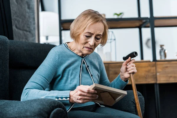 Traurige Seniorin sitzt mit Gehstock und blickt auf Bilderrahmen — Stockfoto