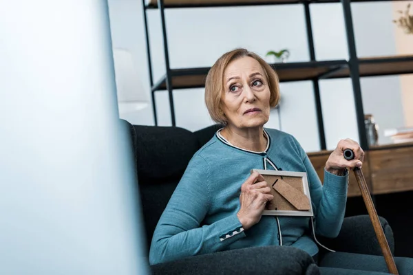 Расстроена старшая женщина сидит с тростью и держит фотографию рамку дома — стоковое фото